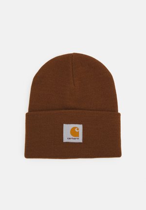Gorro de punto marrón con puño doblado, que presenta una etiqueta blanca cuadrada con un logo amarillo y el nombre de la marca. Textura suave y diseño ajustado.