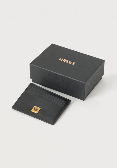 Porte-cartes en cuir noir avec emblème doré Méduse à côté d'une boîte noire avec logo Versace doré sur fond blanc.