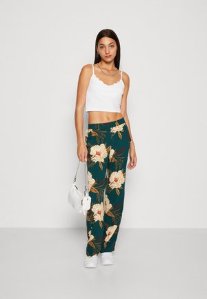White scalloped crop top paired with teal floral trousers featuring large cream flowers. Model holds a small white handbag. White sneakers complete the look.