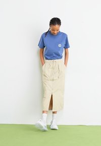 Tricou din bumbac albastru cu buzunar pe piept, asociat cu o fustă midi bej, cu fermoar frontal și talie cu cordon. Pantofi sport albi completează ținuta.
