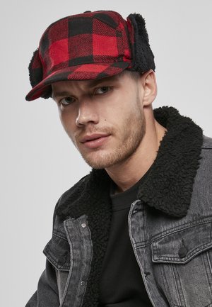 Rot-schwarze karierte Mütze mit Fellfutter und Ohrenklappen, getragen von einer Person in einem schwarzen Shirt und einer grauen Jacke mit Sherpa-Kragen, vor einem hellen Hintergrund.