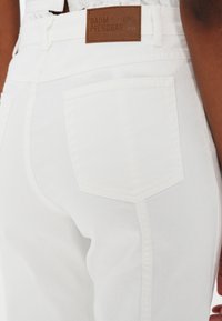 Gros plan sur la poche arrière d'un jean en denim blanc avec un patch en cuir marron portant le logo Baum und Pferdgarten sur la ceinture.