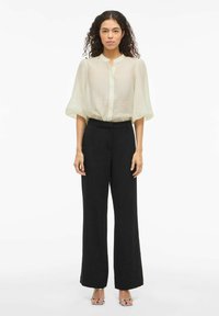 Blusa ligera, vaporosa y color crema con mangas cortas y abullonadas, y botones al frente; combinada con pantalones negros fluidos de cintura alta y sandalias de tacón transparente.