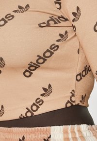 Zblízka na meruňkově zbarvené tričko a šortky Adidas s opakujícími se černými logy Adidas a trojlístkovými vzory na látce.
