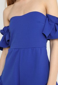 Modrý overall s odhalenými rameny a volánkovými rukávy, vyrobený z hladké tkaniny. Disponuje upnutým živůtkem a volným střihem nohavic.