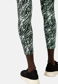 Leggings aderenti in verde e nero con un motivo astratto e una texture liscia, abbinati a scarpe da ginnastica nere con suola testurizzata.