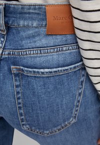 Vaqueros de mezclilla azul claro con una etiqueta de marca de cuero, que cuentan con cinco bolsillos y un sutil desgaste. Camisa a rayas parcialmente visible.