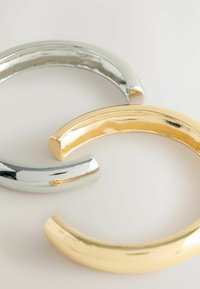 Tre buede armringe i sølv- og guldtone. Den sølvfarvede armring har en skinnende finish, mens den guldtonede har en glat, reflekterende overflade.