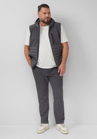 Graue gepolsterte Weste mit Kapuze, mit Reißverschlusstasche, getragen über einem weißen T-Shirt. Kombiniert mit grauen Hosen und hellen Sneakers, lässiges Outdoor-Outfit.