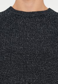 Primer plano de una persona que lleva un suéter de punto texturizado en gris oscuro y negro con un cuello redondo acanalado.