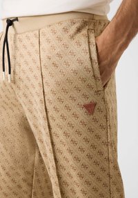 Beige broek met patroon en bruin logo-ontwerp, voorzien van een taille met trekkoord en zijzakken, gemaakt van zacht, gestructureerd materiaal.