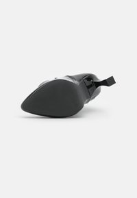 Chaussure à talon haut noire avec une finition brillante, bout pointu, semelle texturée et une petite poignée rectangulaire sur le talon.