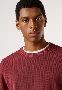 Bordeauxroter, gerippter Strickpullover mit Rundhalsausschnitt, über einem weißen Hemd getragen. Der Stoff hat eine strukturierte Oberfläche und eine legere Passform.