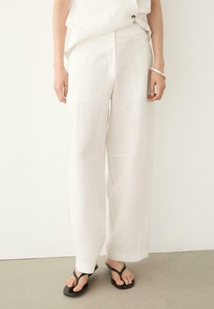 Donna che indossa pantaloni bianchi larghi, top bianco senza maniche, braccialetto d'argento e sandali infradito neri, in piedi contro uno sfondo uniforme.