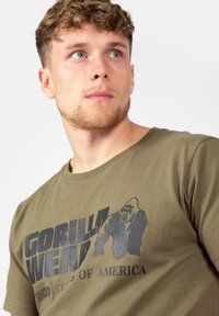 Olivinis žalias medvilninis marškinėliai su dideliu juodu grafiniu spausdinimu gorilos ir tekstu 'GORILLA WEAR UNITED STATES OF AMERICA'.