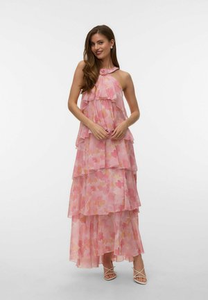 Nő ujjatlan, réteges, rózsaszín virágos maxiruha halter nyakkendős rózsa díszítéssel és fehér pántos magas sarkú cipőben, mosolyogva, összekulcsolt kézzel.