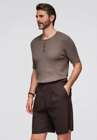 Maglietta corta a righe marrone con maniche corte e tre bottoni, abbinata a pantaloni corti plissettati color marrone scuro a vita alta. Modello mostrato di profilo.