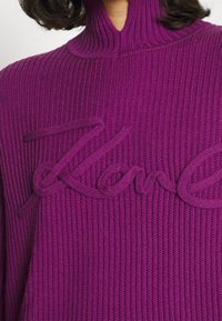 Fioletowy sweter z prążkowanej dzianiny z wysokim kołnierzem oraz wyraźnym teksturowanym logo "Karl" w dopasowanym fioletowym kolorze z przodu.