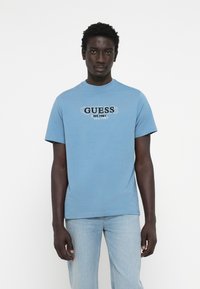 Modrá bavlnená tričko s prednou grafikou, na ktorej je nápis "GUESS" a "EST. 1981" v kontrastných farbách. Krátke rukávy a okrúhly výstrih.