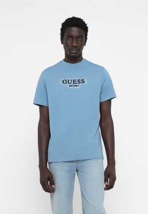 Camiseta de algodón azul con un estampado gráfico frontal con "GUESS" y "EST. 1981" en colores contrastantes. Manga corta y escote redondo.