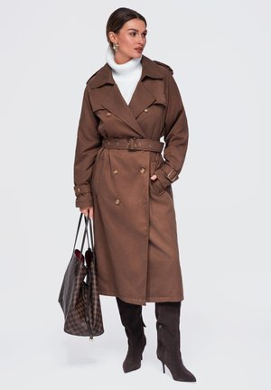Femme portant un trench-coat marron ceinturé, un col roulé blanc, des bottes hautes foncées jusqu'aux genoux, tenant un sac cabas à carreaux, regardant sur le côté.