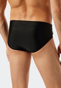 Schwarze Schwimmshorts mit glatter Textur, niedrigem Bund und minimaler Abdeckung. Mit einem dezenten Logo an der Seite.