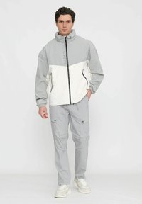 Giacca leggera a blocchi di colore in grigio e bianco, con colletto alto, zip frontale e tasche laterali. Abbinata a pantaloni cargo grigi e sneakers.