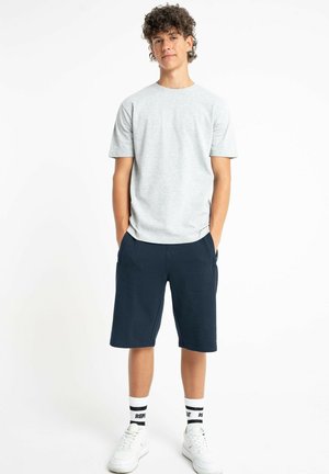 Młody mężczyzna o kręconych włosach, ubrany w jasnoszary t-shirt, granatowe spodenki, białe skarpetki i białe trampki, stoi z rękami w kieszeniach.