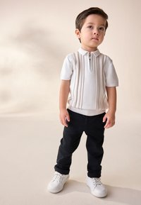 Biela polokošeľa s krátkym rukávom so zipsom a vertikálnymi pásikmi, kombinovaná s čiernymi nohavicami a bielymi teniskami. Stojí na neutrálnom pozadí.