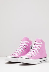 Růžové plátěné vysoké tenisky s bílými tkaničkami, gumovými špičkami a černými akcenty na podrážce. Na boku se nachází kulaté logo.