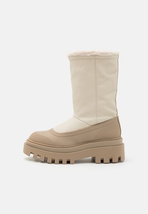 Cizmă din piele bej, cu guler pufos de nuanță chihlimbar, talpă robustă, vârf rotund și accente texturate. Detalii de cusătură verticală vizibile.