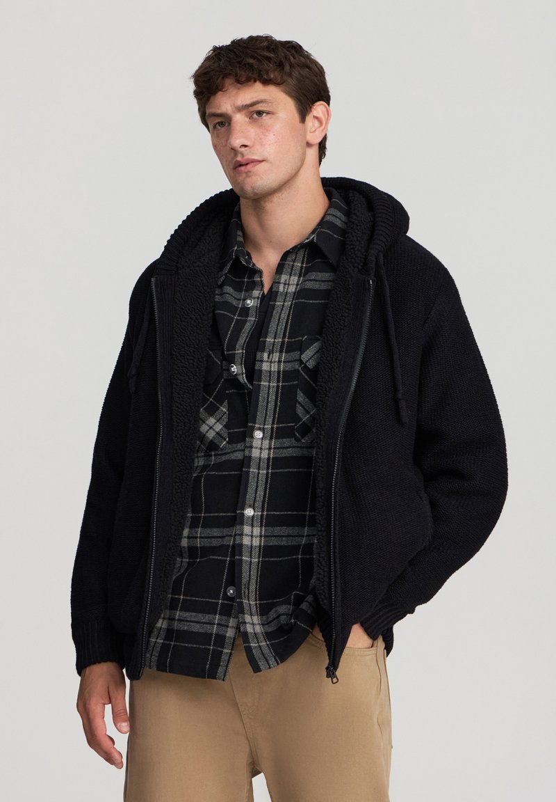 Felpa nera con zip, rivestita internamente in pile testurizzato e dotata di cappuccio regolabile, indossata sopra una camicia a quadri nera e grigia.