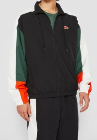 Men's zip-up jacket in black with color-blocked sleeves in green, white, and orange. Features drawstrings, elastic cuffs, and logo detail.