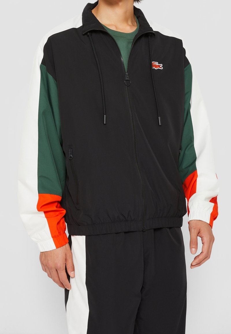 Men's zip-up jacket in black with color-blocked sleeves in green, white, and orange. Features drawstrings, elastic cuffs, and logo detail.