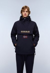 Navy blue anorak with a front pocket, featuring a logo patch and a Norwegian flag. Made of water-resistant material, it has a drawstring hood.