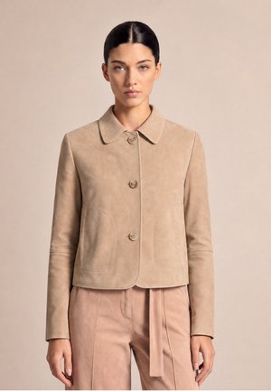 Woman with dark hair wears a beige buttoned jacket and matching high-waisted pants against a plain beige background.
