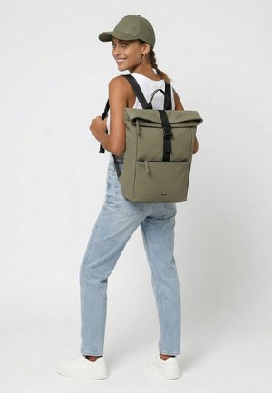 Vrouw met een groene pet, wit hemdje, lichtblauwe spijkerbroek, witte sneakers en een olijfgroene rugzak met zwarte banden, glimlachend en omkijkend.