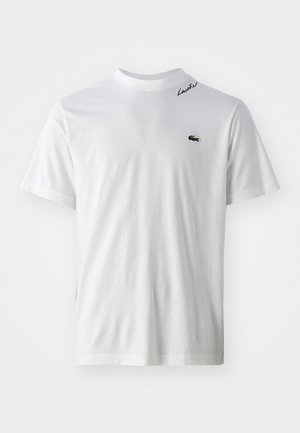 T-shirt bianco a maniche corte con piccolo logo di coccodrillo verde sul lato sinistro del petto e testo "Lacoste" nero vicino al collo sul lato destro.