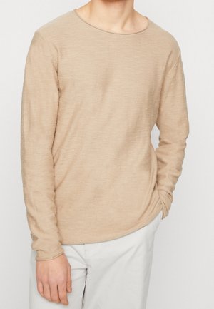 Beige Langarmpullover mit rundem Halsausschnitt, strukturiertem Stoff und enganliegenden Ärmeln, kombiniert mit hellen Hosen.