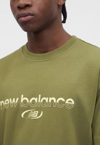 Άνδρας που φοράει πράσινο ελιάς T-shirt New Balance με λευκό λογότυπο και κείμενο, καδράρισμα που δείχνει τον λαιμό και το πάνω μέρος του στήθους.