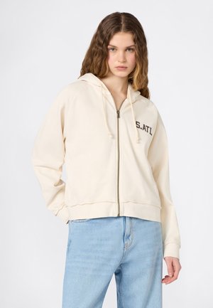 Joven con cabello ondulado que lleva una sudadera con cremallera color crema con el texto "S.ATL" y jeans azul claro, posando frente a un fondo blanco liso.