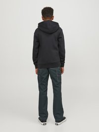 Haut à capuche noir, associé à un pantalon vert foncé avec des coutures visibles et des poches cargo. Des baskets noires et blanches complètent le look.