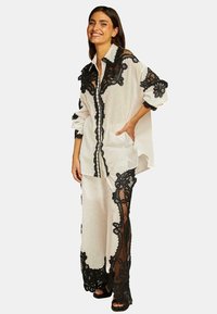 Completo a due pezzi composto da una camicia bianca e pantaloni decorati con intricati dettagli in pizzo nero. Maniche lunghe e vestibilità comoda.