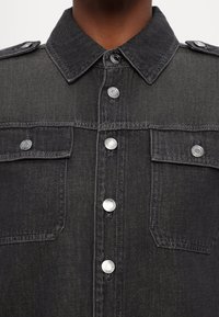 Gros plan d'une personne portant une veste en jean gris foncé avec des boutons-pression argentés et deux poches poitrine avec rabats à boutons.
