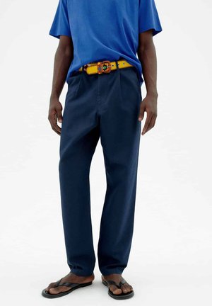 A person wearing a blue shirt, navy pants, a yellow patterned belt, and black flip-flops standing against a plain background.