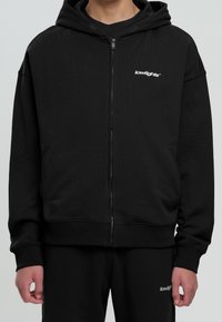 Zwarte zip-up hoodie van gestructureerde stof met een ruime capuchon, voorzakken en een wit logo op de linker borst. Heeft ook ribgebreide manchetten.