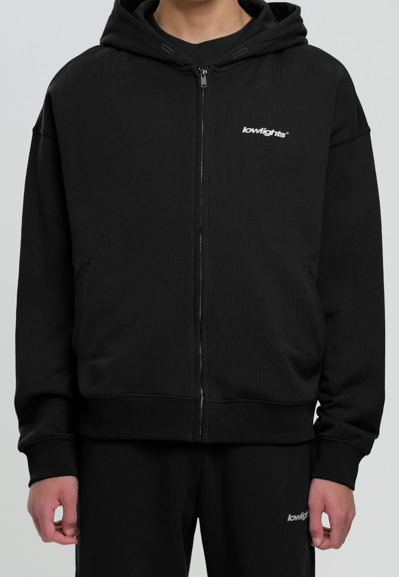 Zwarte zip-up hoodie van gestructureerde stof met een ruime capuchon, voorzakken en een wit logo op de linker borst. Heeft ook ribgebreide manchetten.