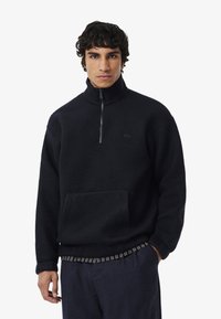 Pullover in pile nero con colletto alto e zip a metà, caratterizzato da una tasca frontale e un logo discreto. Tessuto testurizzato con una vestibilità morbida.