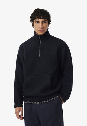Black fleece pullover with a high collar and half-zip, featuring a front pocket and subtle logo. Textured fabric with a relaxed fit.