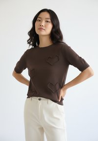 Brązowy sweter z krótkim rękawem z trzema teksturowanymi wzorami serc na przodzie, zestawiony z jasnymi spodniami. Gładka dzianina.